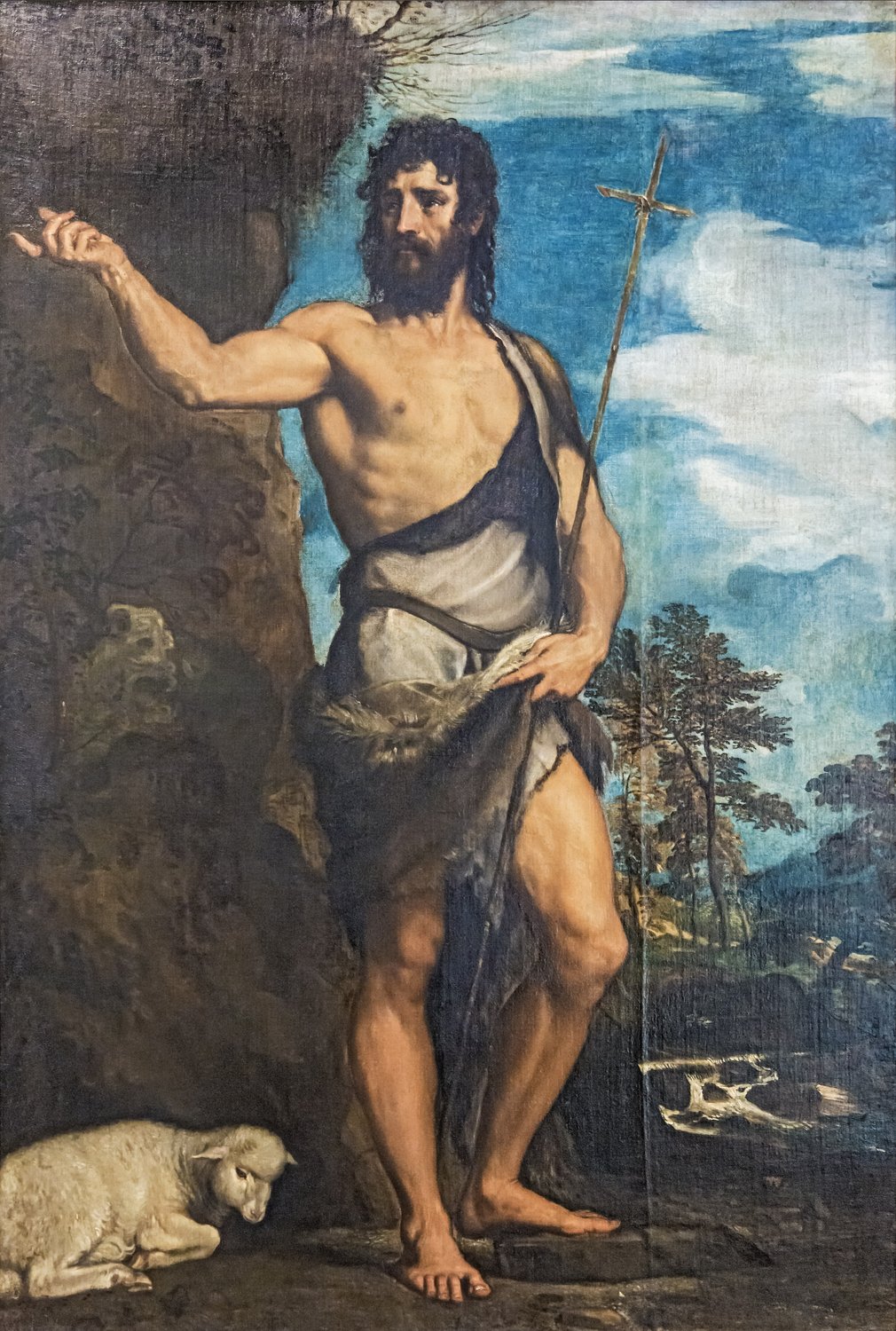 A painting by Titian: John the Baptist stands in the wilderness, a strong man with dark curling hair and a thick black beard, bare but for a camel skin and a white cloth about his waist. He points up and away with his right hand and holds a tall, thin cross of reed in his left. A lamb lies at his feet by a rock, and a stream and trees lie behind him under a blue sky.
