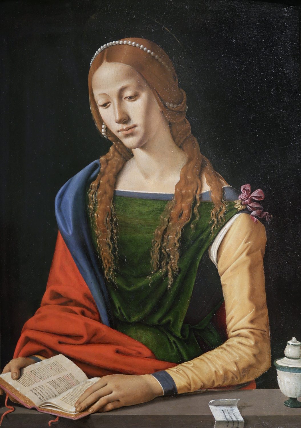 A painting by Piero di Cosimo: Mary Magdalene, a young woman with long wavy auburn hair threaded with pearls, sits at a stone ledge and looks down at a small open book. She wears a green dress with pale yellow sleeves and a red cloak lined with blue. A white lidded jar stands at the right, against a dark background.