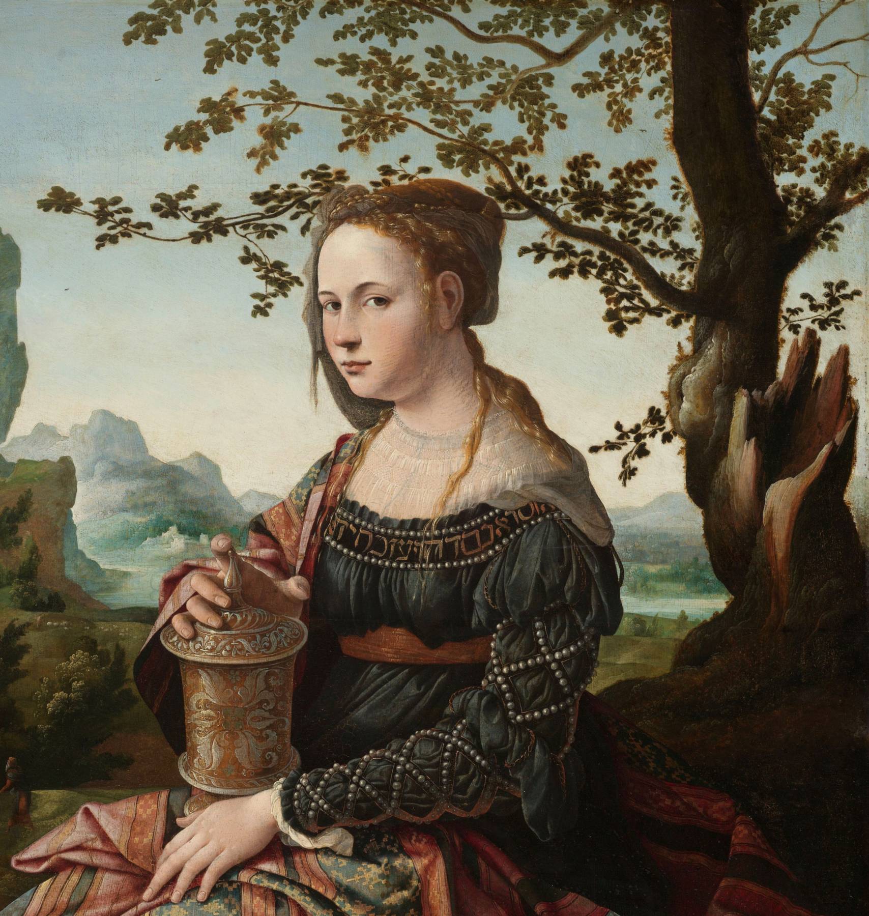 A painting by Jan van Scorel: Mary Magdalene, a young woman with golden-red hair braided and pinned up under a thin grey veil, sits beneath a tree and looks calmly out. On her lap she holds a tall golden jar of ointment. She wears a dark gown sewn with rows of pearls, its neckline lettered in gold, and a striped shawl. Behind her, pale rocky crags rise over a blue-green valley.