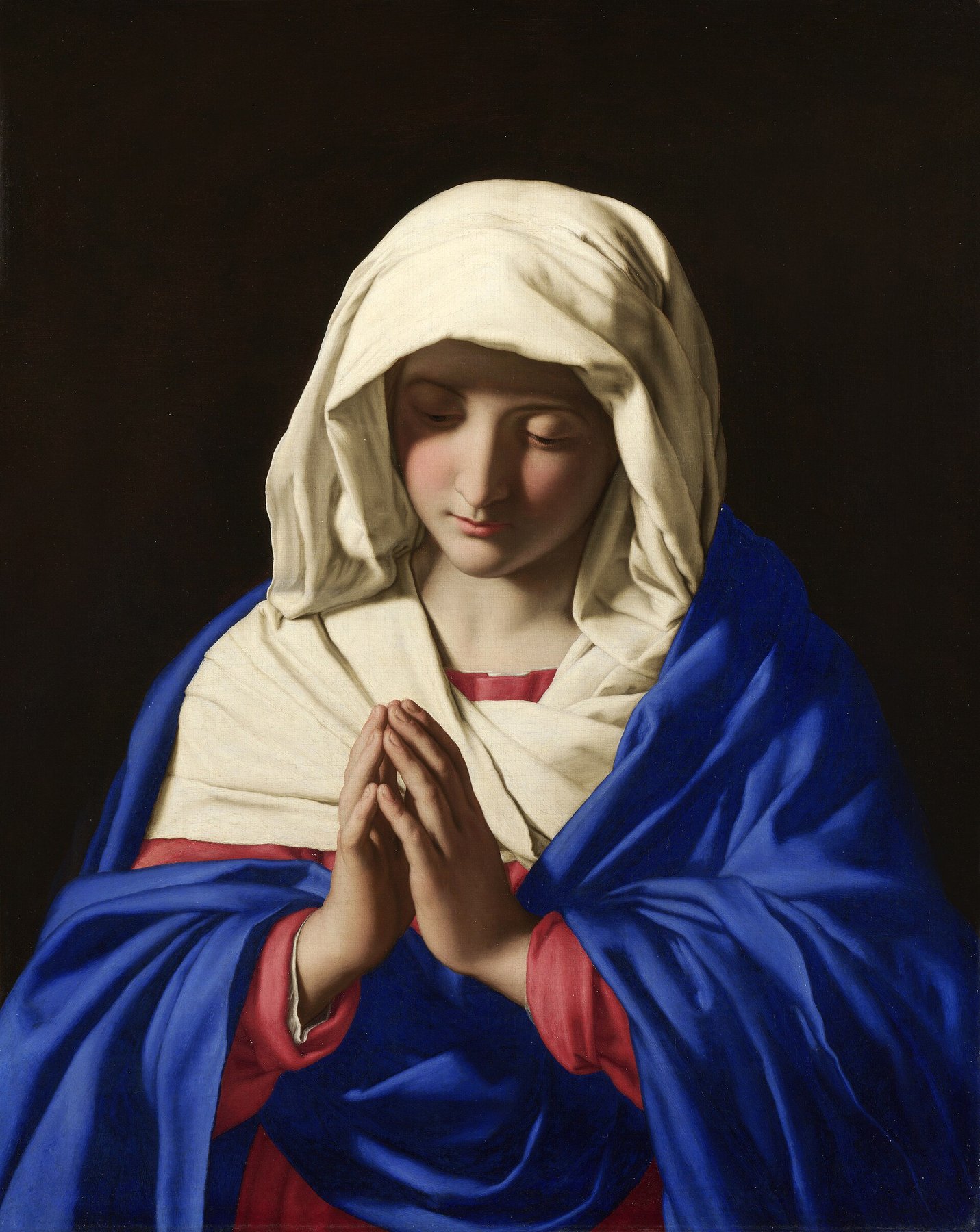 A painting by Sassoferrato: Mary, a young woman with lowered eyes, her hands joined in prayer. A white veil covers her head. She wears a red dress under a deep blue mantle, against a dark background.