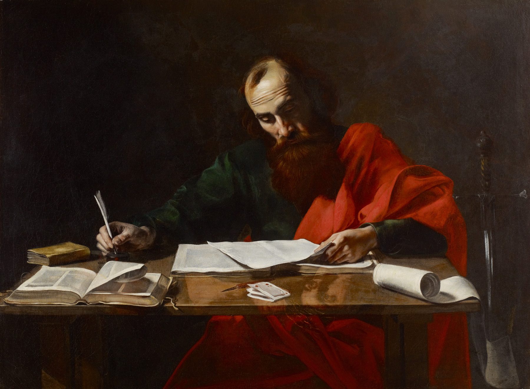 A painting: Paul, balding, with a long red-brown beard, sits at a wooden table writing with a quill, his eyes on the page. He wears a dark green tunic and a bright red cloak. Open books, loose sheets and a scroll lie on the table, and the hilt of a sword stands at the right edge.