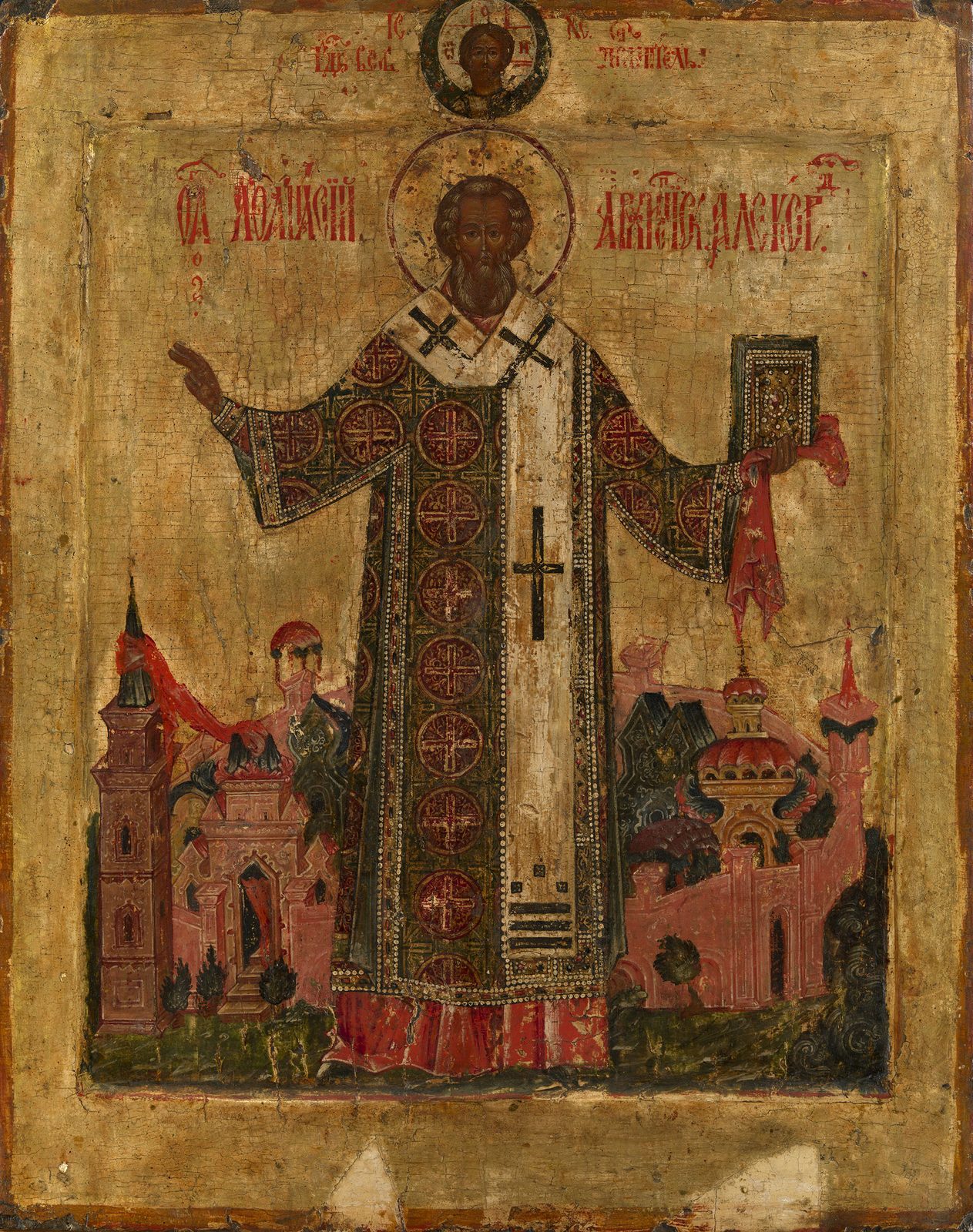 A Russian icon: Athanasius stands full length in a patterned vestment, blessing with his right hand and holding a jewelled book in his left, before a town of red towers and onion domes, with Christ in a small medallion above his halo