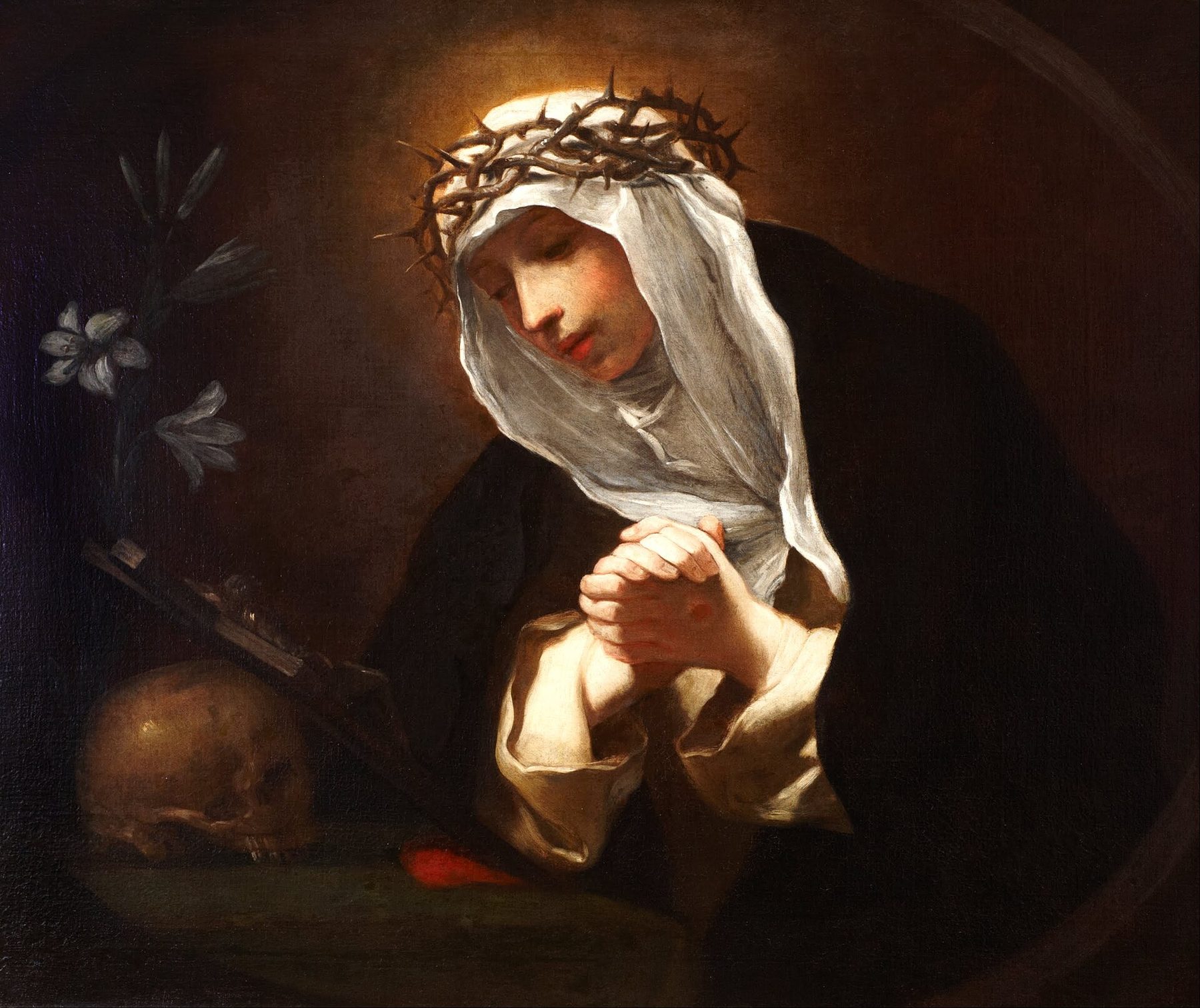 A painting: Catherine of Siena in a white veil and black Dominican mantle, with a crown of thorns on her head, eyes lowered and hands clasped in prayer. A lily and an open book stand beside a skull on the table at the left, against a dark background.
