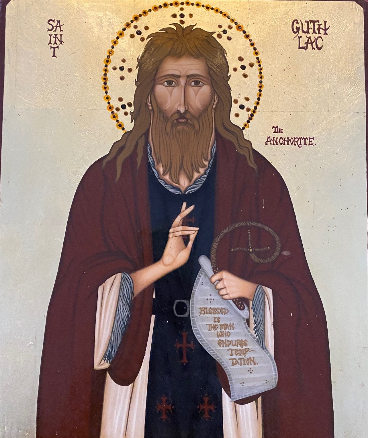 A modern icon on gold: Guthlac, with long brown hair, a long beard and a halo, in a dark red cloak over a dark robe, raises his right hand and holds a scroll reading "Blessed is the man who endures temptation"; the words Saint Guthlac the Anchorite are painted beside him