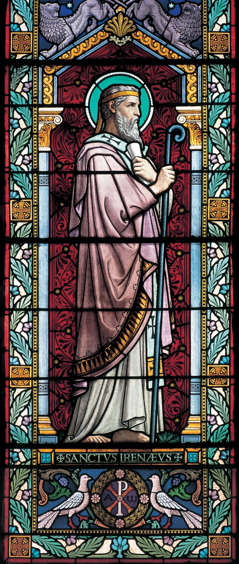 A stained-glass window: Saint Irenaeus in profile, an old bearded bishop with a green halo and a gold band in his hair, holding a scroll and a tall blue crozier, in a pink cloak before a deep red patterned ground, with SANCTVS IRENÆVS beneath him and two doves by a Chi-Rho below