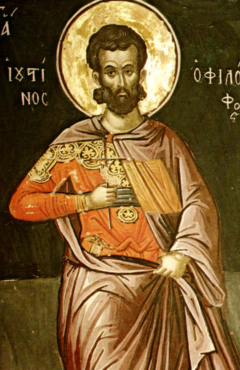 A fresco of Justin as a dark-haired, bearded man with a gold halo, in a red tunic and a pale cloak, holding a small cross and a closed book, with his name in Greek beside him