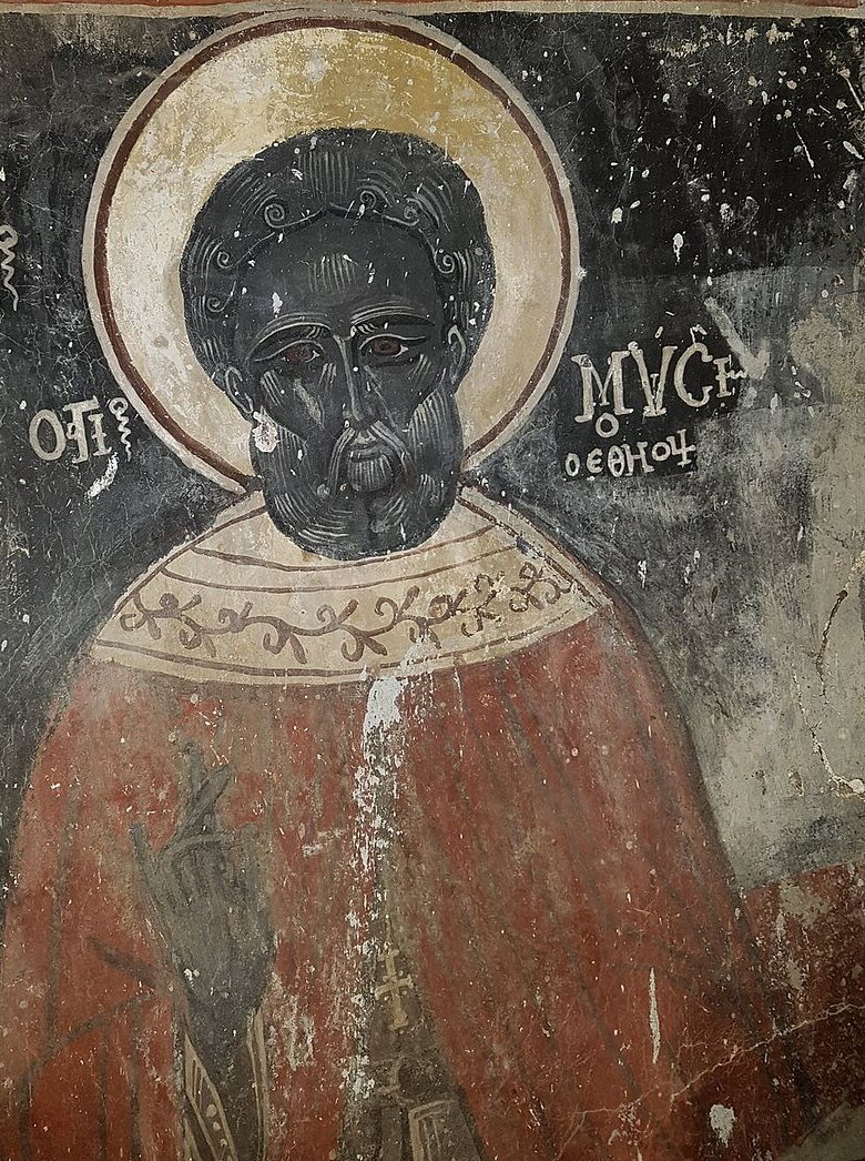 A worn wall painting: Moses as a black man with a dark beard and a round head of dark hair, inside a pale gold halo on a black ground. He wears a red cloak with an embroidered collar and raises one hand. Greek letters beside his head name him.