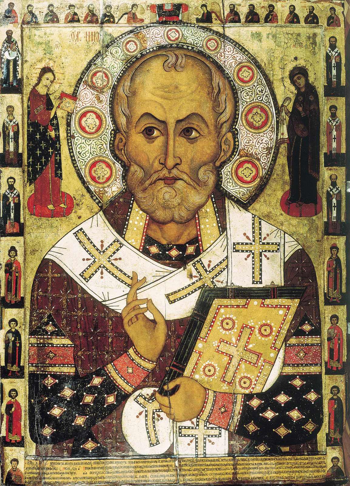 A Russian icon of 1294: Saint Nicholas, bald with a curling forelock, large dark eyes and a full white beard, in a white vestment with black crosses. He blesses with one hand and holds a jewelled Gospel book, before a round halo set with red medallions. Small figures of Christ and the Mother of God stand by his shoulders, and rows of small saints fill the border.
