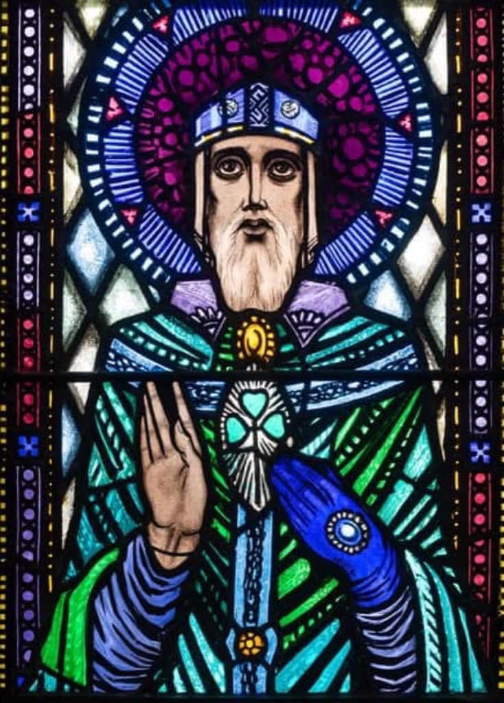 A stained-glass window in deep blues, greens and purples: Saint Patrick, with a long white beard and wide dark eyes under a jewelled band, raises his right hand in blessing, a green shamrock clasped at his breast, a great purple halo behind his head