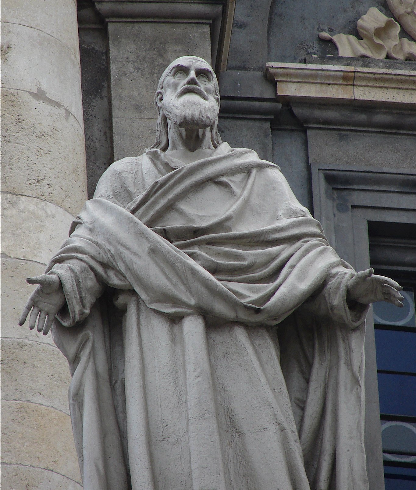 A pale stone statue of Polycarp as an old bearded man in a heavy draped cloak, arms held out low with open hands, face lifted toward the sky, against the grey stone and a dark window of a church wall