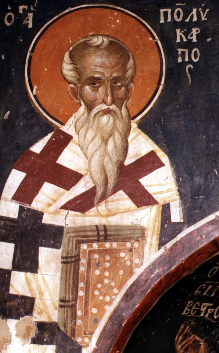 A Byzantine fresco: Saint Polycarp, an old bishop with a high forehead, tired dark eyes and a long pointed white beard, before an orange halo on a deep blue wall. He wears a white vestment with large crosses and holds a jewelled Gospel book. His name is in Greek letters beside his head.